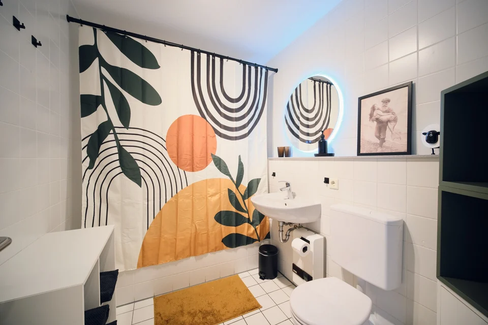 Badezimmer mit Badewanne, farbigem Duschvorhang, Waschbecken, rundem Spiegel und WC. – Apartment Mittendrin.