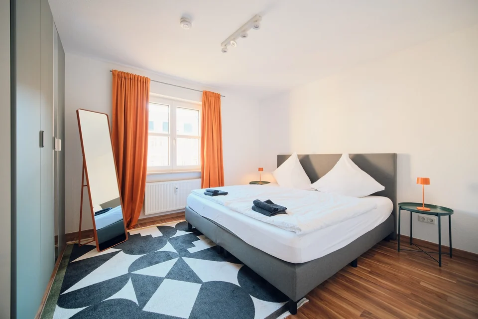 Doppelbett, Nachttische und Standspiegel im Schlafzimmer mit orangefarbenen Vorhängen. – Apartment Mittendrin.